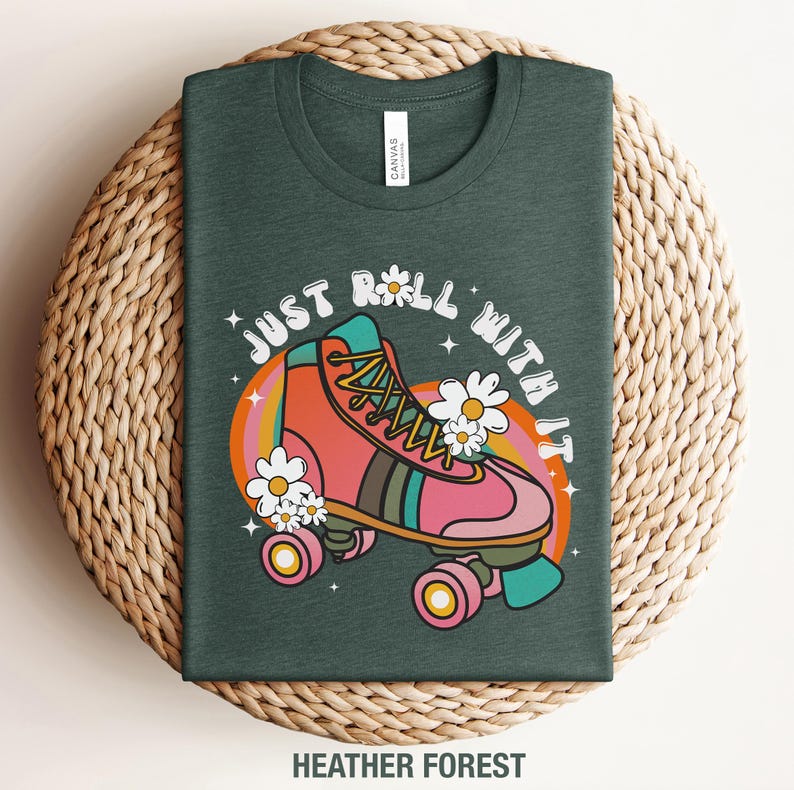 May include: A heather forest green t-shirt featuring a graphic of a pink roller skate with yellow laces, surrounded by white daisies and a colorful rainbow arch. The text "JUST ROLL WITH IT" is printed in a white arched font above the skate.