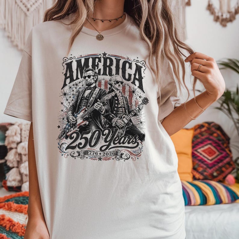 May include: Beige t-shirt featuring a graphic with the word "AMERICA" above an illustration of two figures playing guitars. The graphic also includes "250 Years" and the dates "1776-2026".