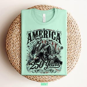 May include: Mint green t-shirt featuring a graphic of two historical figures playing guitars, with the word "AMERICA" above them. The design includes the text "250 Years" and "1776-2026". The shirt is displayed on a woven surface.