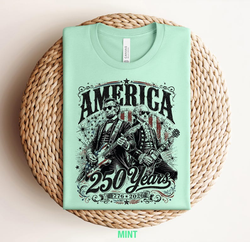 May include: Mint green t-shirt featuring a graphic of two historical figures playing guitars, with the word "AMERICA" above them. The design includes the text "250 Years" and "1776-2026". The shirt is displayed on a woven surface.