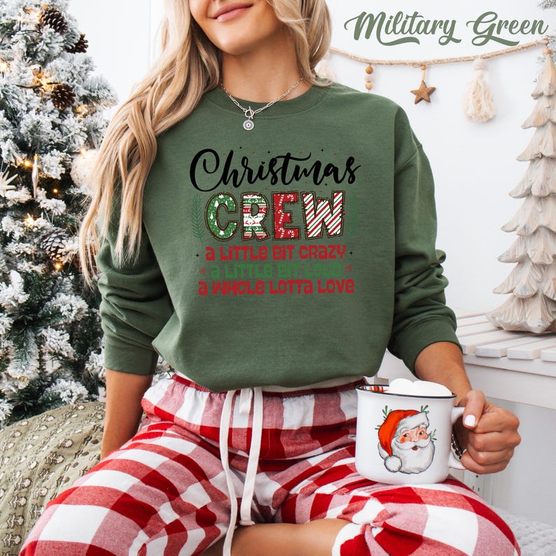 May include: A military green sweatshirt with the text "Christmas CREW" in black, red, and green. The sweatshirt also has the phrases "a little bit crazy", "a little bit fun", and "a whole lotta love" in red. A white mug with a Santa Claus graphic is held in the hands.