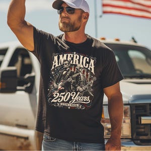 May include: Dark gray t-shirt with the word "AMERICA" above an image of two historical figures playing guitars. The graphic includes the text "250 Years" and the years "1776-2026". The shirt is worn by a person wearing sunglasses and a white cap.