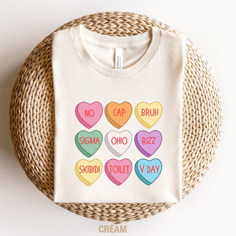 May include: A cream colored t-shirt with a design of nine candy hearts with different phrases on them. The phrases are "NO", "CAP", "BRUH", "SIGMA", "OHIO", "RIZZ", "SKIBIDI", "TOILET", and "V DAY".