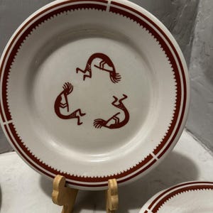 May include: A white ceramic plate with a red border and a design of three stylized figures in a circle. The figures are brown and have a Native American design.