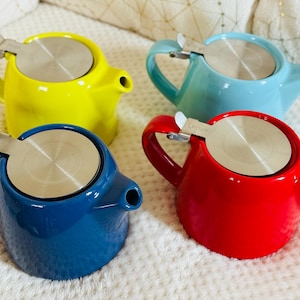 Puede incluir: Cuatro pequeñas teteras de colores en amarillo, azul, rojo y turquesa. Cada tetera tiene una tapa y un asa de acero inoxidable. Las teteras están sobre una superficie blanca texturizada.