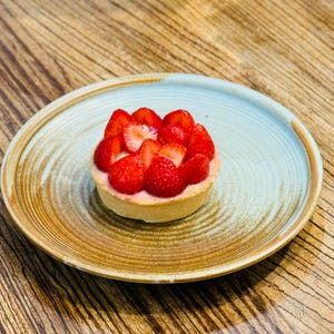 Puede incluir: Una tarta de fresas sobre un plato de cerámica con borde azul claro y borde marrón. La tarta presenta una base de masa rellena de crema rosa y cubierta con fresas frescas en rodajas. El plato está sobre una superficie de madera.