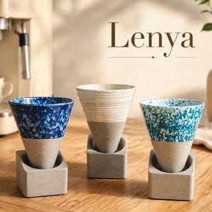 Puede incluir: Tres tazas de cerámica para café en azul, blanco y verde azulado, cada una con un diseño moteado único. Las tazas se apoyan en soportes de hormigón gris. La palabra "Lenya" se muestra en marrón.