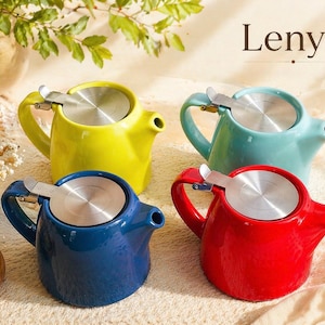 Puede incluir: Cuatro teteras de cerámica coloridas en amarillo, turquesa, azul y rojo. Cada tetera tiene una tapa plateada y un asa curva. La palabra "Lenya" se muestra en la esquina superior derecha.