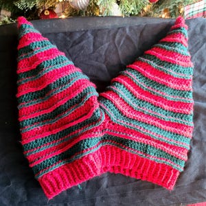 Puede incluir: Un gorro de elfo de ganchillo a rayas rojas y verdes hecho a mano. El gorro presenta dos puntas puntiagudas y está hecho con filas alternas de hilo rojo y verde. El gorro es probablemente para ocasiones festivas.