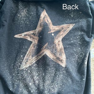 May include: Black hooded sweatshirt with a large, distressed star design on the back. The star is brown with a textured appearance, and there are white paint splatters around it. The word "Back" is printed in white.