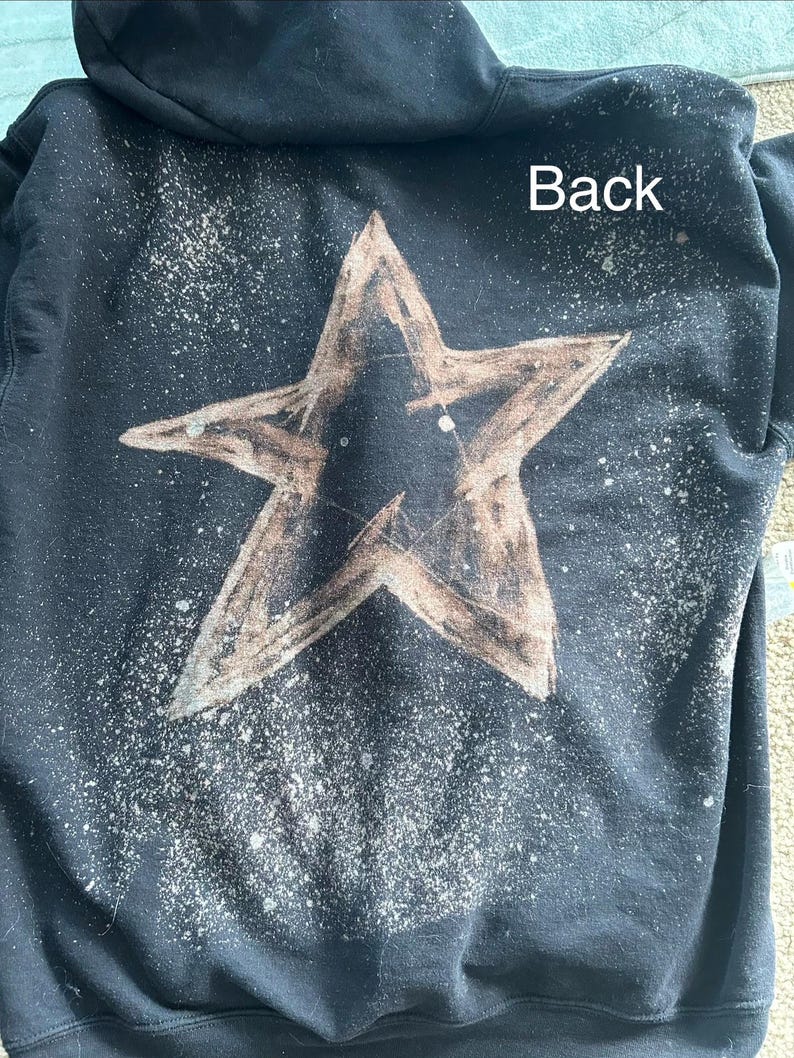 May include: Black hooded sweatshirt with a large, distressed star design on the back. The star is brown with a textured appearance, and there are white paint splatters around it. The word "Back" is printed in white.