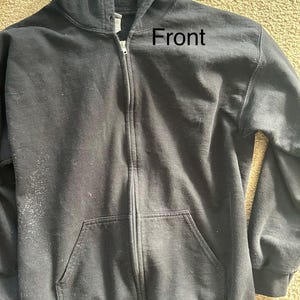 May include: A black zip-up hooded sweatshirt with a front pocket. The word "Front" is printed on the upper right side of the sweatshirt. The sweatshirt has a hood and a full-length zipper.