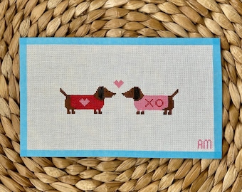 May include: A rectangular needlepoint canvas featuring two brown dachshunds facing each other. The left dog wears a red sweater with a pink heart, and the right dog wears a pink sweater with the letters XO. A small pink heart sits between them.