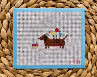 May include: A needlepoint canvas featuring a brown dachshund wearing a colorful party hat, holding three balloons in red, yellow, and blue. A small birthday cake with three candles sits nearby. The letters AM are stitched in pink in the corner.