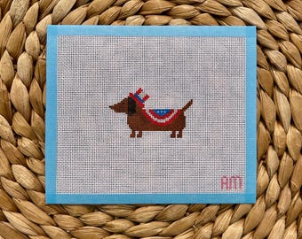 May include: A square needlepoint canvas featuring a brown dachshund wearing a red, white, and blue patterned hat and blanket. The canvas has a light blue border and the letters AM stitched in pink in the bottom right corner.