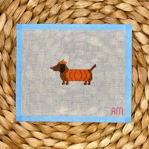 May include: A cross-stitch design featuring a dachshund wearing a pumpkin costume and a pumpkin hat. The artwork is set against a white background with a light blue border. The initials "am" are in the lower right corner.