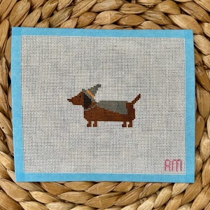 May include: A needlepoint canvas with a blue border featuring a dachshund wearing a witch hat and costume. The canvas has the word "Kim" stitched in pink in the bottom right corner.