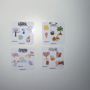 May include: Four white square cards with seasonal illustrations. "Winter" card shows a snowman, tree, and penguin. "Summer" card has a sun, ice cream, and palm tree. "Spring" card features a bunny and flowers. "Autumn" card shows a squirrel and owls.