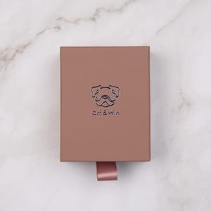 May include: A rectangular, light brown box with a dog illustration and the text "arf & win" in a minimalist style. A pink ribbon is attached to the box. The box is on a white marble surface.