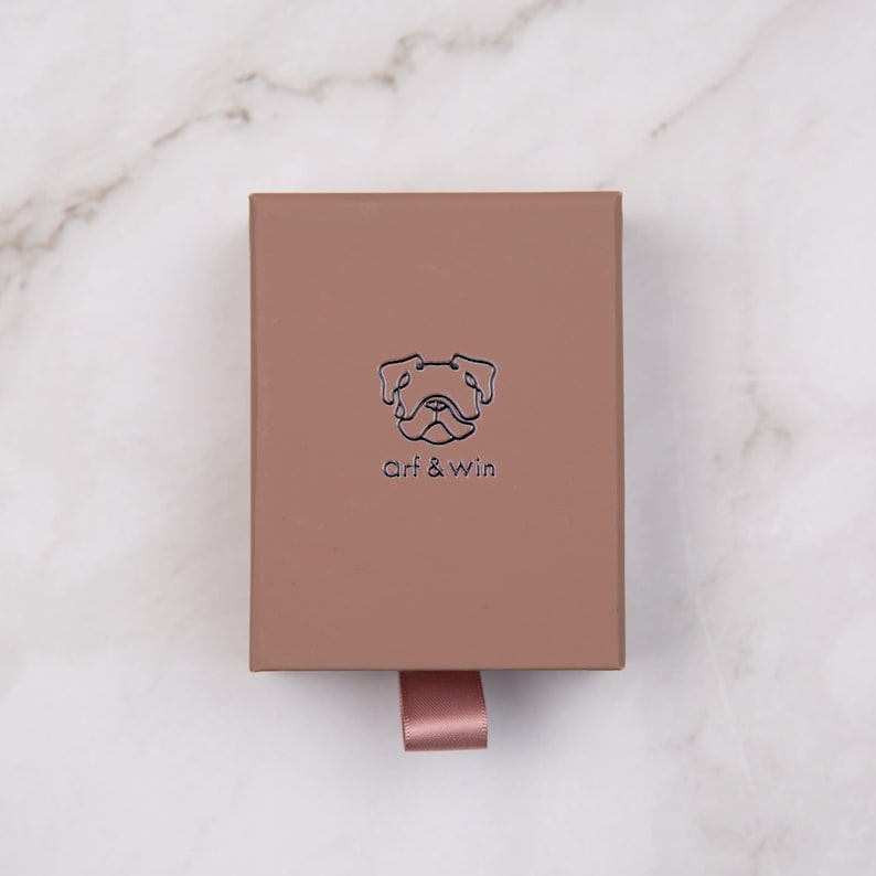 May include: A rectangular, light brown box with a dog illustration and the text "arf & win" in a minimalist style. A pink ribbon is attached to the box. The box is on a white marble surface.