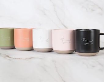 May include: A set of five ceramic mugs in olive green, peach, white, light pink, and black. Each mug features a simple white line drawing of a dachshund dog on the front and has a natural, unglazed tan base.