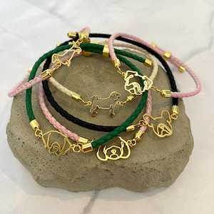 May include: A collection of braided bracelets in black, green, pink, and cream colors. Each bracelet features a gold-toned metal charm shaped like a dog silhouette or face, connected by small gold-toned metal cylinders to the braided cord.