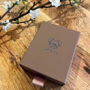 May include: A brown box with the text "Arf & Win" and a bulldog logo. The box has a pink ribbon pull and is on a wooden surface. White flowers and branches are in the background.