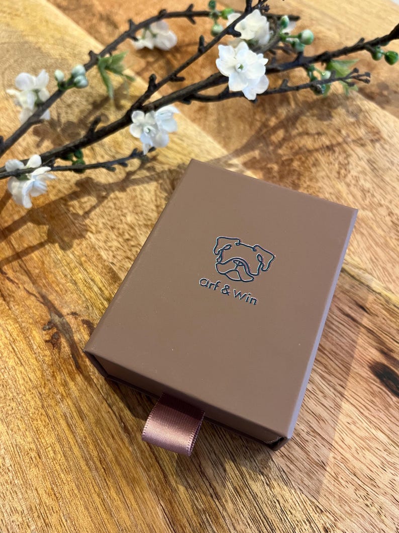 May include: A brown box with the text "Arf & Win" and a bulldog logo. The box has a pink ribbon pull and is on a wooden surface. White flowers and branches are in the background.