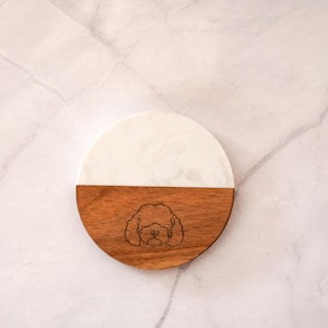 May include: A round coaster with a two-tone design. The top half is white, and the bottom half is wood-toned with a dog face etched into it. The coaster is on a marble surface.