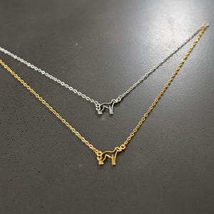 May include: Two delicate necklaces, one gold-toned and one silver-toned, each featuring a small dog silhouette pendant. The pendants are connected to fine chains. The necklaces are displayed against a dark background.