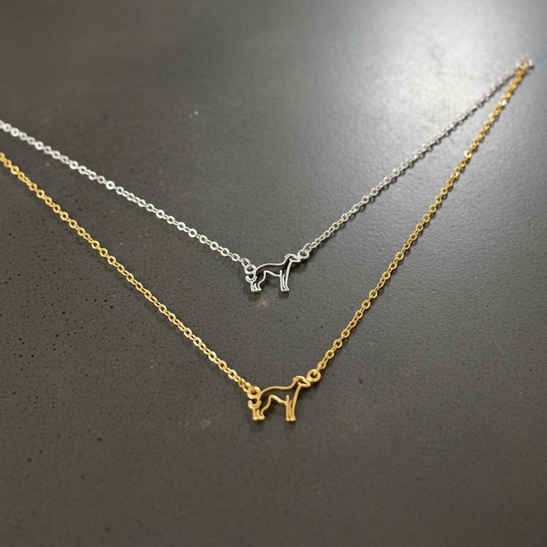 May include: Two delicate necklaces, one gold-toned and one silver-toned, each featuring a small dog silhouette pendant. The pendants are connected to fine chains. The necklaces are displayed against a dark background.