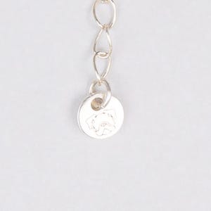 May include: A silver-colored necklace chain with a small, round pendant. The pendant features a detailed engraving of a dog's face. The chain is composed of small, interconnected links, and the pendant is attached at the end.