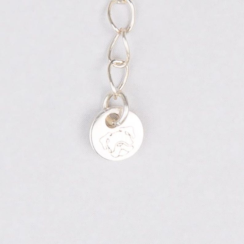 May include: A silver-colored necklace chain with a small, round pendant. The pendant features a detailed engraving of a dog's face. The chain is composed of small, interconnected links, and the pendant is attached at the end.