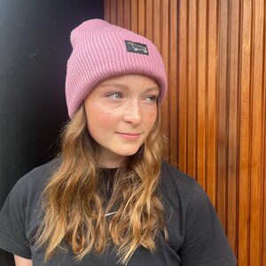 Puede incluir: Un gorro de punto acanalado rosa con un pequeño parche rectangular con la silueta de un perro salchicha negro. El gorro es de color liso y tiene un borde doblado. El fondo es una pared de madera.