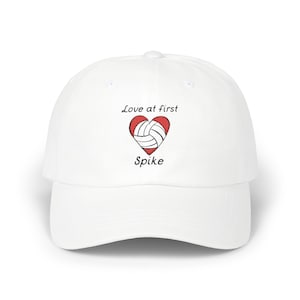 Puede incluir: Gorra de béisbol blanca con el texto "Love at first Spike" y un corazón rojo con un balón de voleibol dentro.