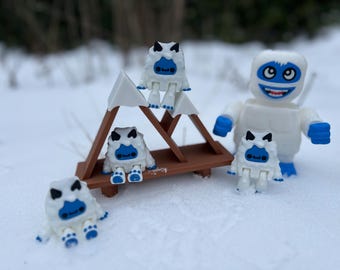 May include: Four white and blue yeti figurines, two sitting on a wooden mountain-shaped stand and two standing on a snowy surface. One larger yeti figurine is standing on the snowy surface.