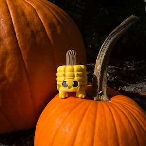 May include: Two large orange pumpkins with textured surfaces. A small, yellow corn-shaped figurine with black eyes and a brown husk sits atop one pumpkin, creating a playful autumn scene. The pumpkins are in natural sunlight.