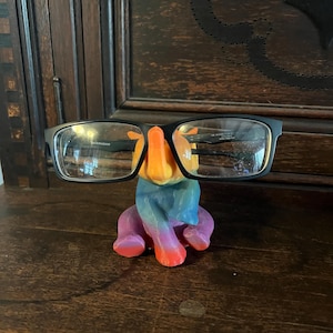 May include: A black-framed pair of eyeglasses rests on a colorful, hand-painted elephant-shaped glasses holder. The elephant is painted with shades of orange, blue, purple, and red. The glasses holder is sitting on a dark wooden surface.