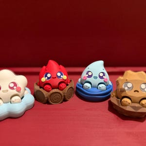 May include: Four colorful, cartoonish figurines representing elements: a cloud, fire, water droplet, and rock. Each has large eyes and is set on a base. The cloud is white and blue, the fire is red, the water droplet is light blue, and the rock is tan.
