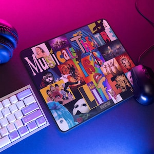Op de afbeelding: Een kleurrijk muismatje met een collage van afbeeldingen en de tekst "Musical Theatre is Life". Het muismatje heeft een zwarte rand en ligt op een roze en blauwe ondergrond.