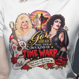 May include: A white t-shirt featuring a graphic print of Dolly Parton with an acoustic guitar and Dr. Frank-N-Furter. The text reads: Go ahead, make your own kind of time warp. Legends never really leave us... they just find a bigger stage.