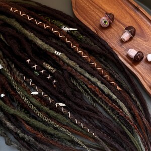 May include: A collection of brown, green, and auburn dreadlocks, adorned with silver and copper-colored decorative wraps and charms. A wooden tray holds mushroom-shaped beads. The dreadlocks are arranged on a dark green surface.
