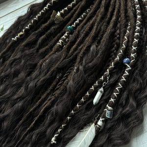 May include: Dark brown synthetic dreadlocks with white stitching and various charms, including a feather, a crystal, and a butterfly.