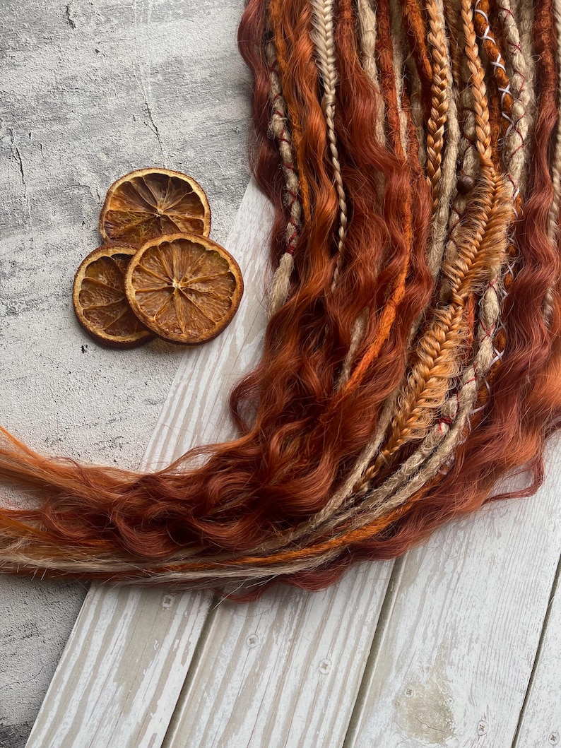 May include: A set of brown, blonde, and orange synthetic dreadlocks with a wavy texture. The dreadlocks are arranged in a loose braid and are lying on a white wooden surface.