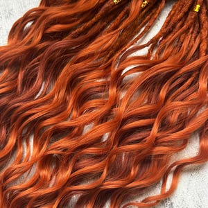 May include: Close-up of vibrant orange and auburn dreadlocks with wavy ends. The synthetic hair extensions have a natural look, with some strands featuring small gold-colored beads. The dreadlocks are arranged on a light gray surface.
