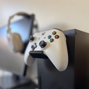May include: A white Xbox controller with black buttons and colored action buttons is mounted on a black console. The controller has a sleek design and is positioned to be easily accessible. The background is blurred, suggesting a focus on the gaming setup.