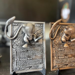 May include: Two 3D printed Pokemon cards featuring Mew and a bronze-colored card with a different Pokemon. The cards are on black stands and have detailed artwork and text. The text on the silver card reads "Mew 2 70HP Basic Mimicry Rainbow Wave".