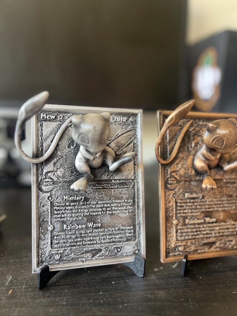 May include: Two 3D printed Pokemon cards featuring Mew and a bronze-colored card with a different Pokemon. The cards are on black stands and have detailed artwork and text. The text on the silver card reads "Mew 2 70HP Basic Mimicry Rainbow Wave".