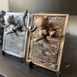May include: A 3D printed bronze Pokemon card featuring a  Eevee with the text "Mimicry" and "Rainbow Wave".