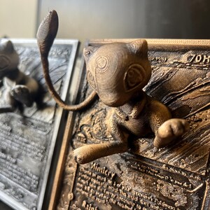 May include: A 3D printed bronze-colored Pokemon card featuring a detailed sculpture of the Pokemon, Mewtwo, with its signature psychic powers. The card is titled "Rainbow Wave" and includes the text "Mimicry".